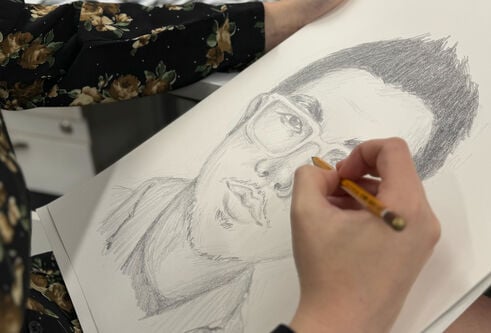 Close up of an unseen individual's hands sketching a face of a man with a pencil