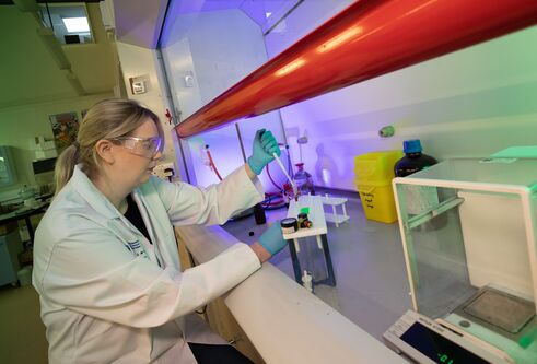 Woman wearing white lab coat, sat at a table wearing gloves and protective safety glasses, using science equipment on a bench