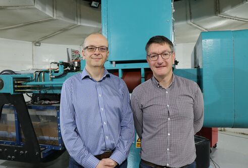 two men wearing checked shirts and glasses, looking and smiling at camera, with large machinery behind them