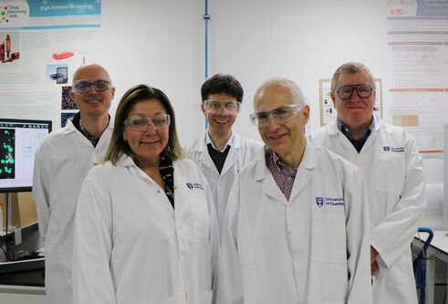 five scientists wearing lab coats and safety glasses, standing next to computer and lab equipment, looking and smiling at the camera