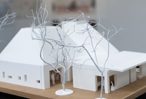 miniature model of a large house made from white material. Tiny little figures stand nearby and there are small branches outside to resemble trees in the winter without their leaves