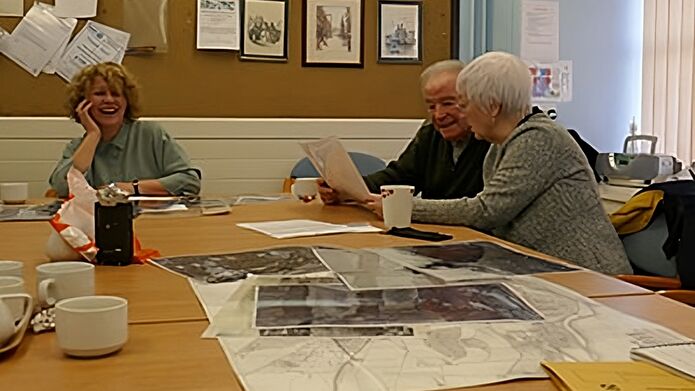 A female archivist and two older community members sit at a table discussing large photograph images of Dundee