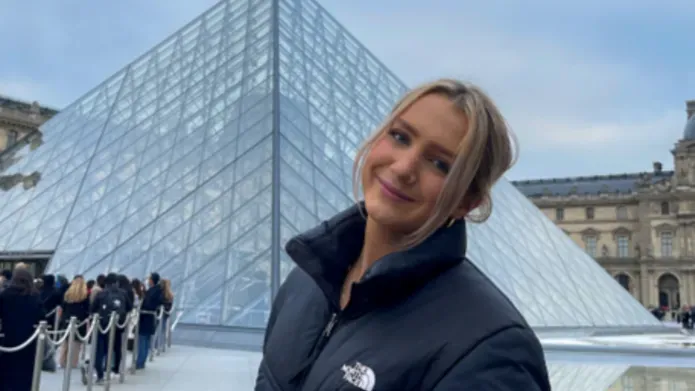 A blonde woman in a black coat is in front of the louvre.