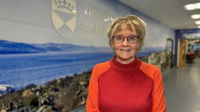 Professor Jill Belch, in a red jumper, standing in front of a blue background with the University of Dundee branding