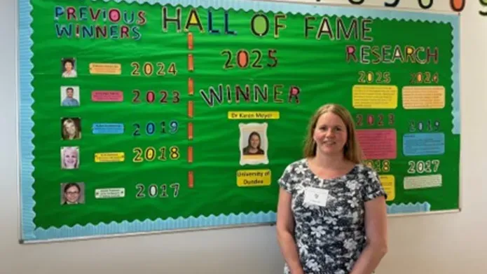 Karen Meyer, 2025 winner, stands in front of a Mathematics Hall of Fame 2025 poster 