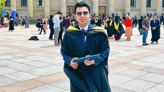 A man in sunglasses and graduation robes stands in front of a grand building, others in graduation robes are in the background.
