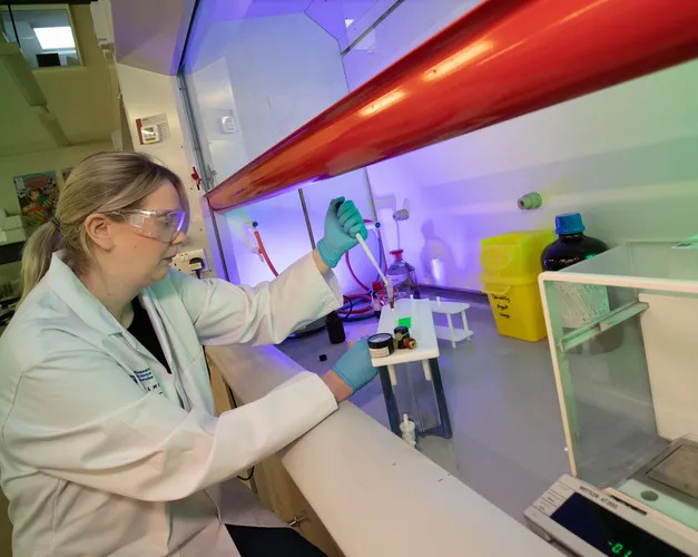 Woman wearing white lab coat, sat at a table wearing gloves and protective safety glasses, using science equipment on a bench