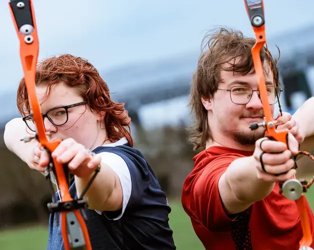 two students standing back to back, each holding an archery bow and pointing it forwards