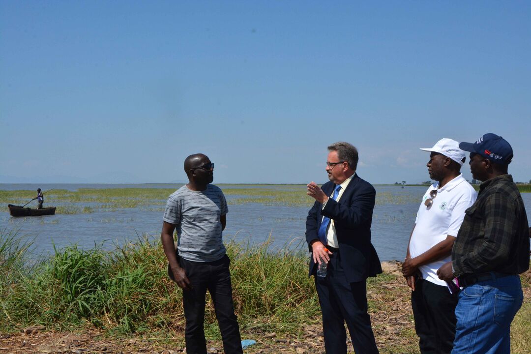 Professor Gillespie standing on the bank of a lake, speaking to three men. There are fishing boats further back in the lake itself.