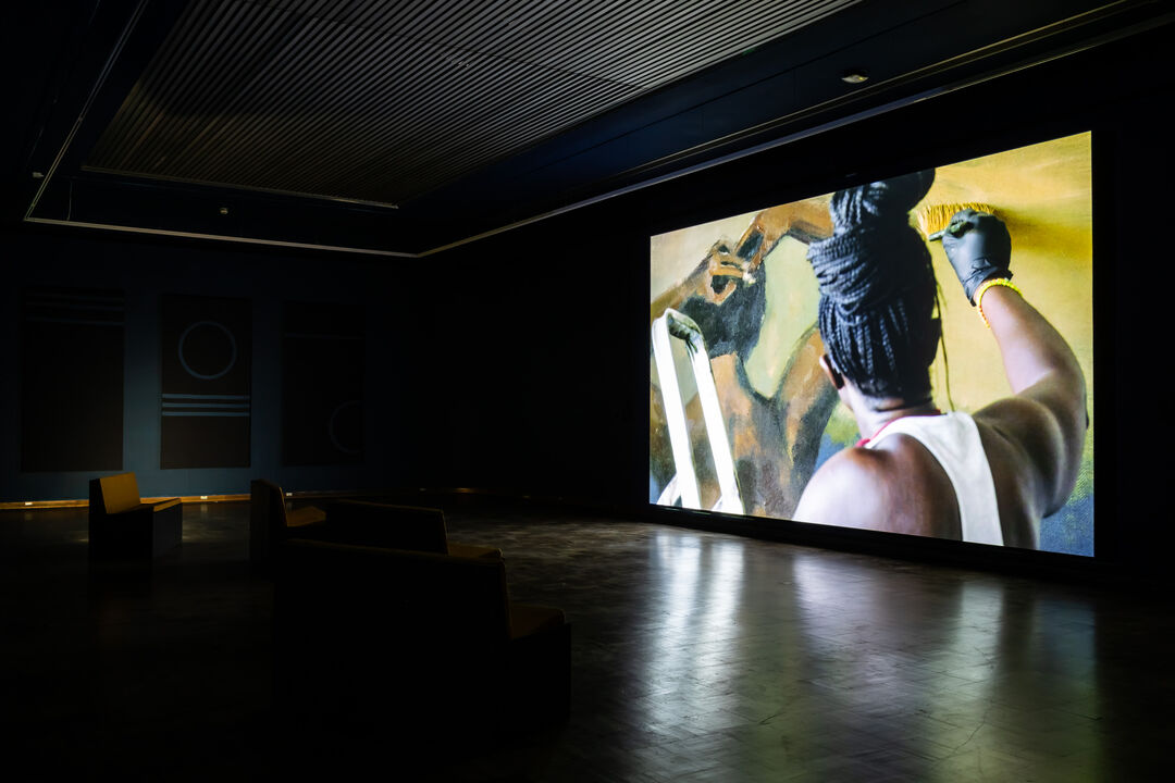 Exhibition view. Projection in a dark gallery shows a close up of a painter at work from behind
