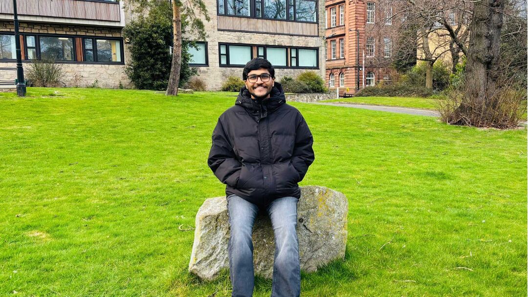 A person sits on a large boulder with their hands in their jacket pockets. There is a building in the background.