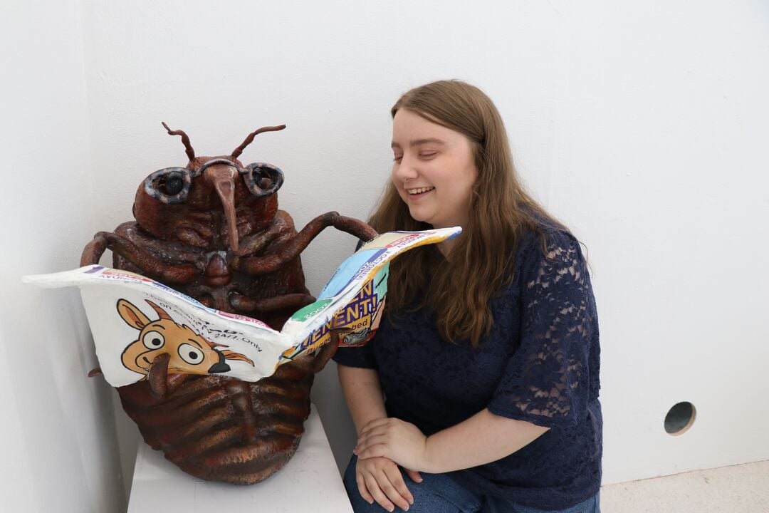 Large artistic creation of a bedbug, wearing glasses and reading the TV listings. Artist peers over the bugs shoulder, looking at the magazine and smiling 