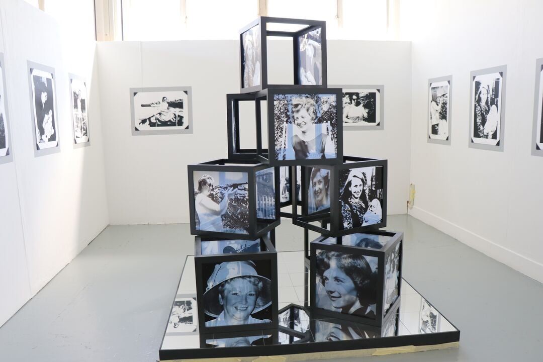 lots of hollow cubes made from dark wooden frames piled on top of each other in a pyramid form. Some cubes feature transparent prints of older women, while others are empty  