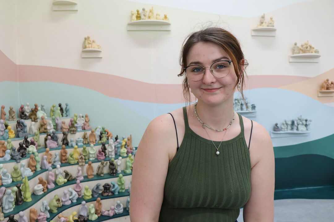 Head and shoulders of young woman with short dark hair and glasses, looking towards the camera and smiling. Behind her are a group of little ceramic figures