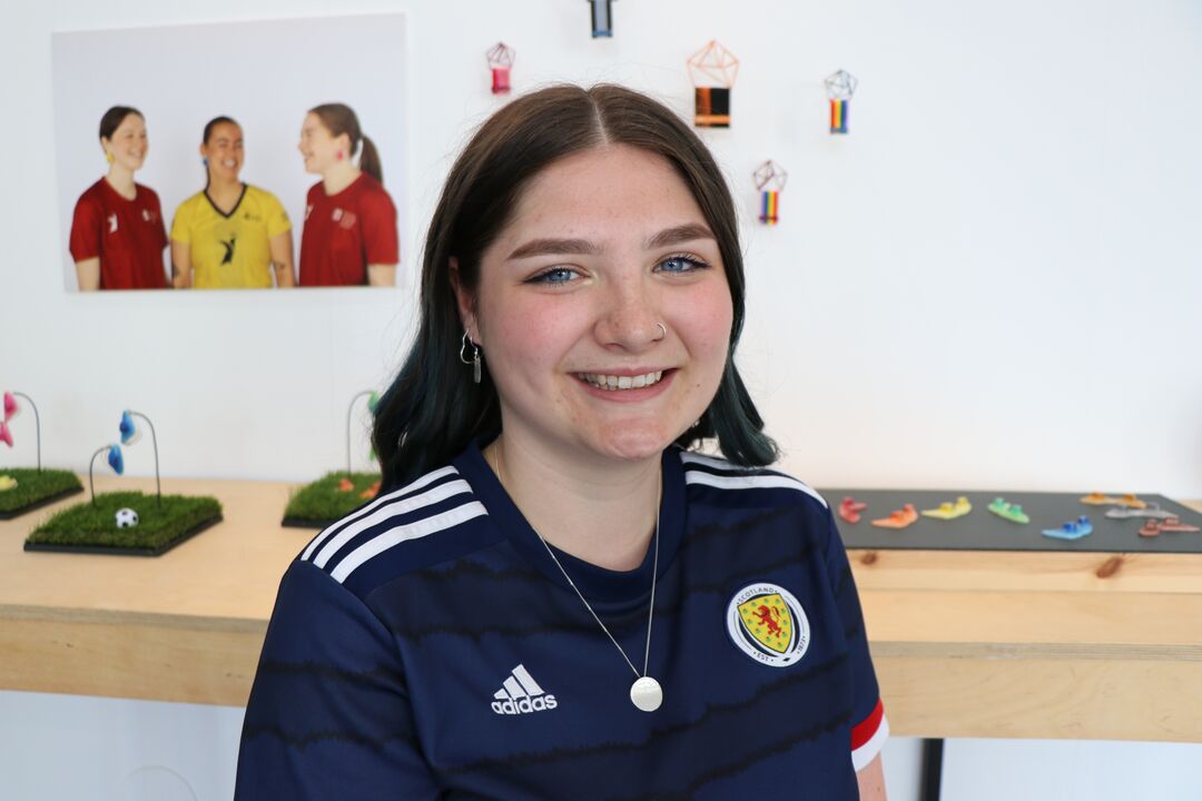 Young woman with long dark hair wearing a Scotland football top, smiling at the camera. A shelf with jewellery is behind her