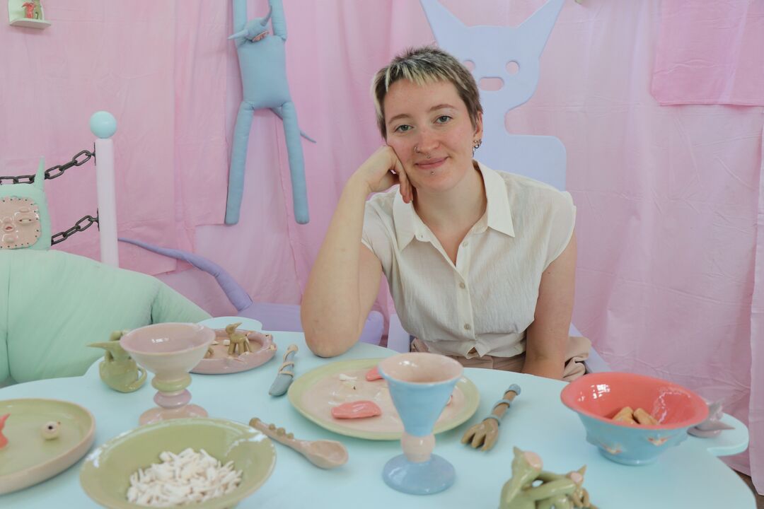 Wound woman sits at a dining table with pastel coloured themed furniture and settings. All settings are ceramic, including bowls full of teeth and plates full of eyeballs 