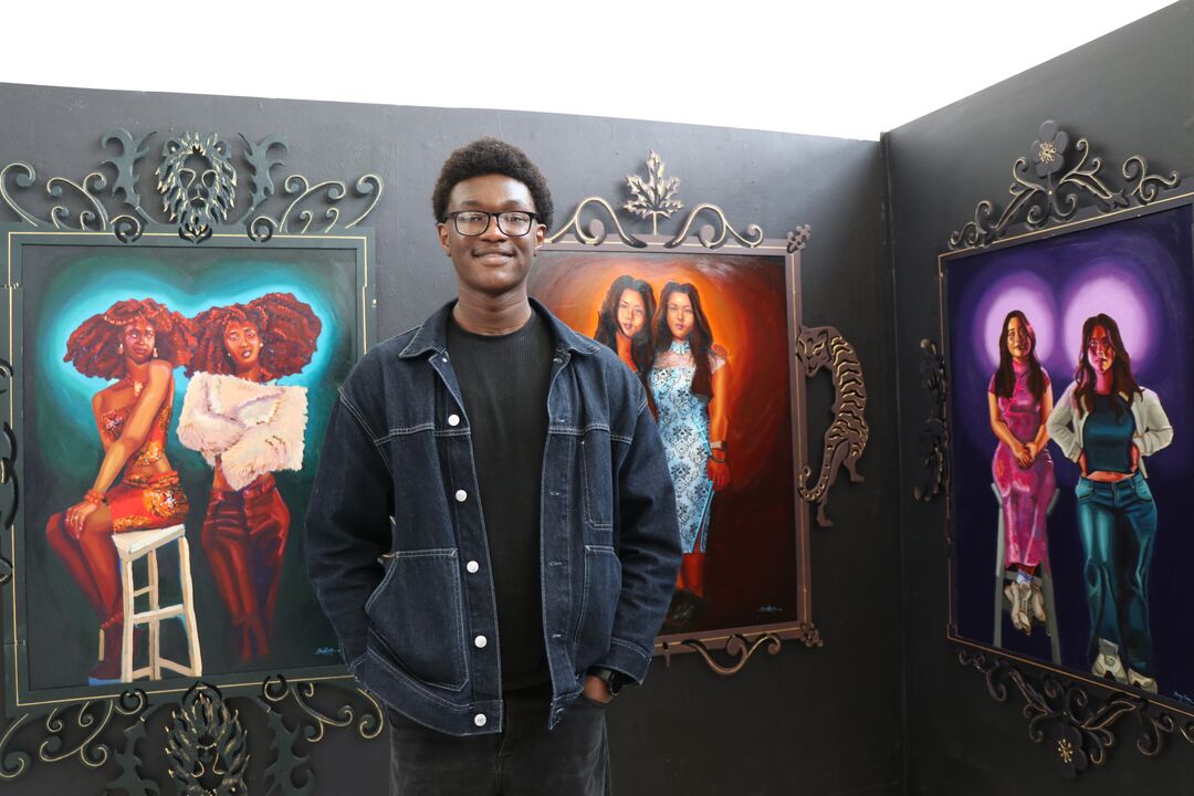 Man wearing a denim jacket and glasses, stands in front of three large oil paintings, smiling