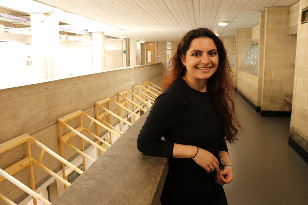 Young woman with long dark hair leaning on an indoor mezzanine edge, looking over to camera and smiling 