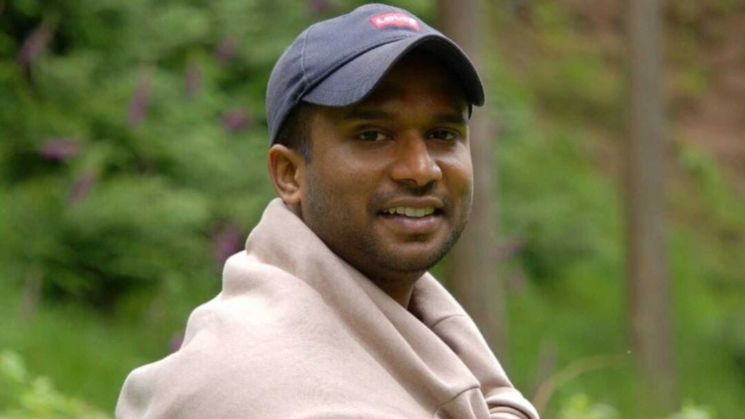 A person wearing a baseball cap stands outside, there is greenery in the background.