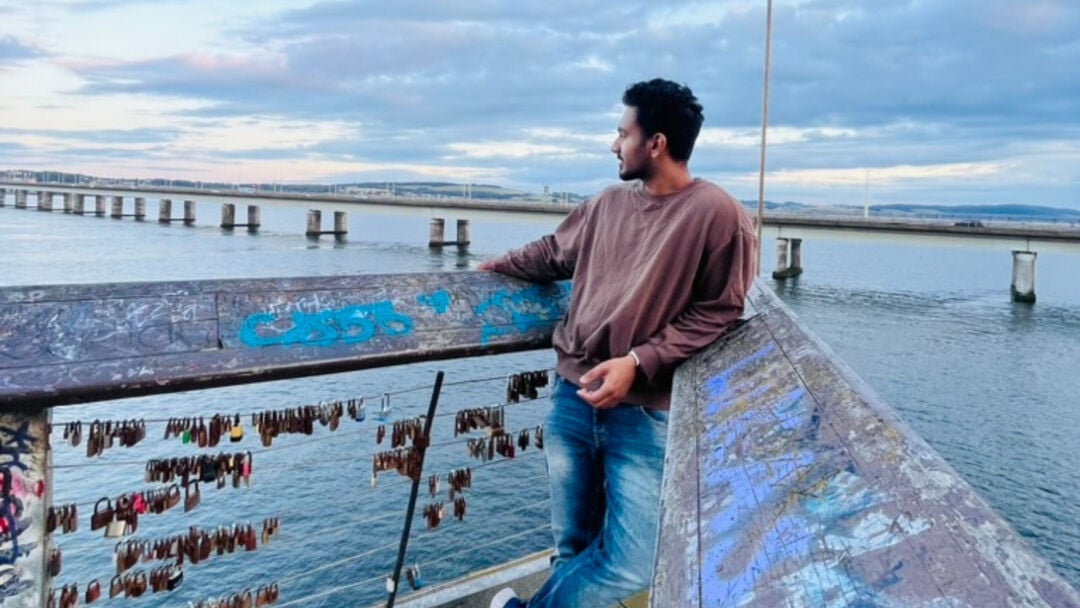 A student stands on a bridge, resting beside a padlock attached to the railing, with the water in the background