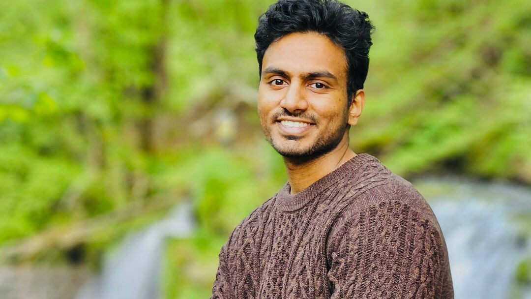 The student smiles at the camera, wearing a brown jumper with a waterfall and greenery in the background