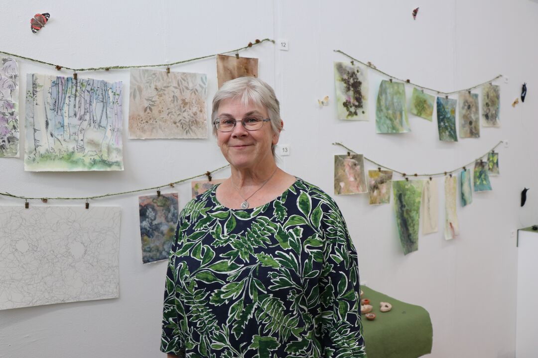 Older woman wearing top with a green leaf pattern on it stands in front of a string of watercolour paintings