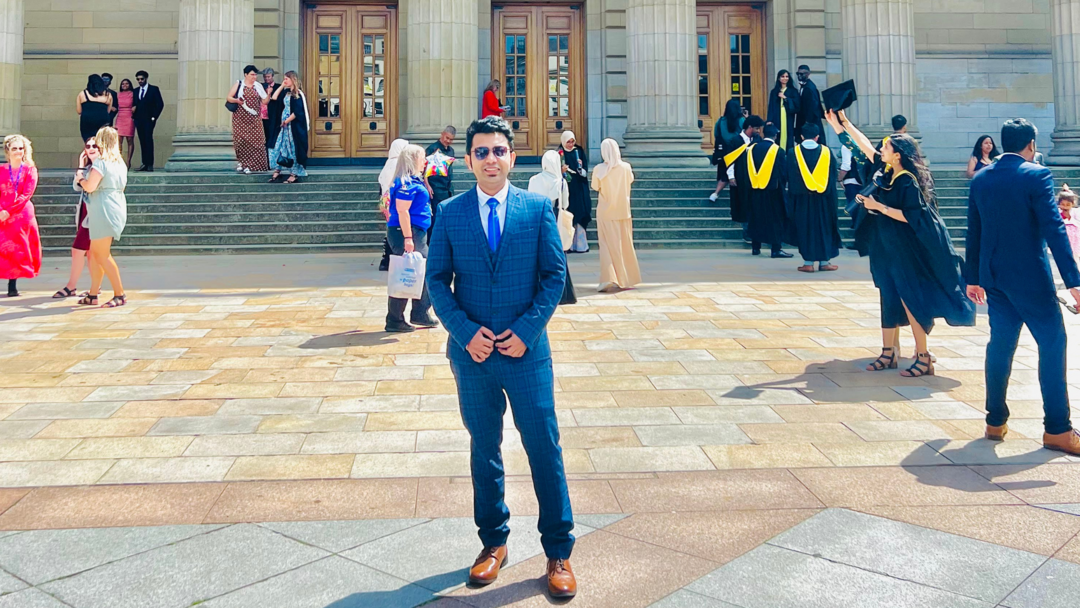 A man dressed in a suit and wearing sunglasses, stands in front of a grand building.