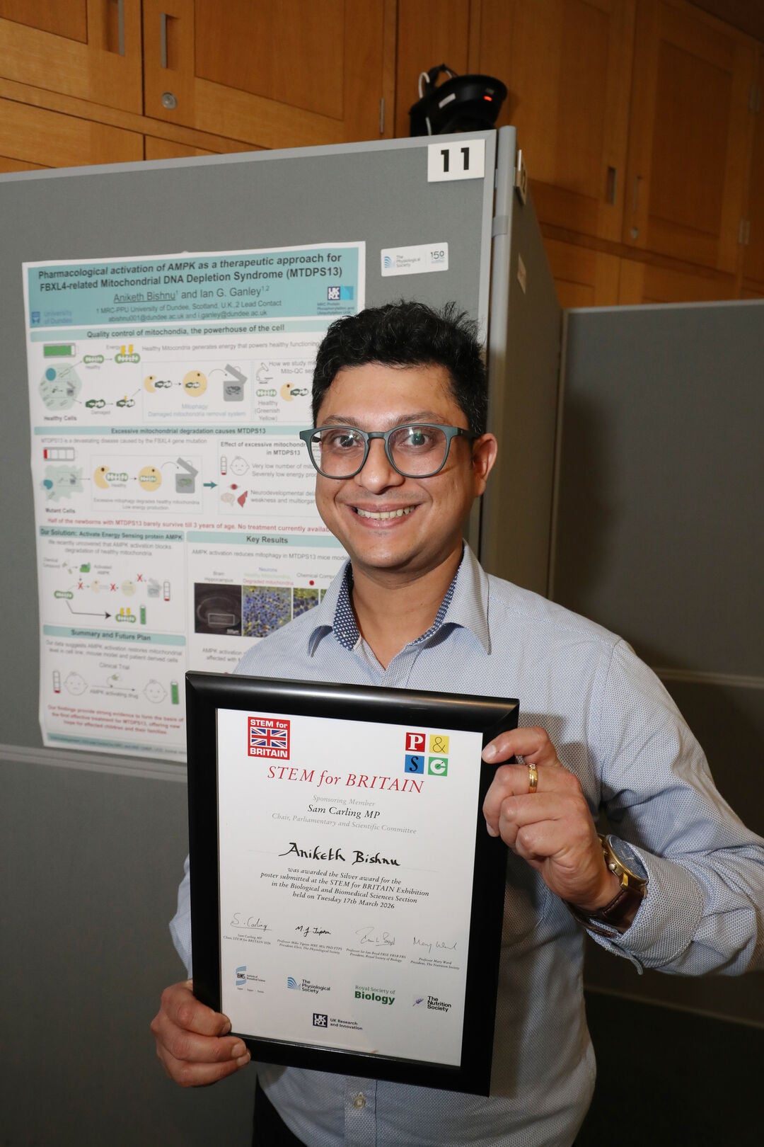 A person stood smiling holding a framed certificate. A poster on a display board is in the background.