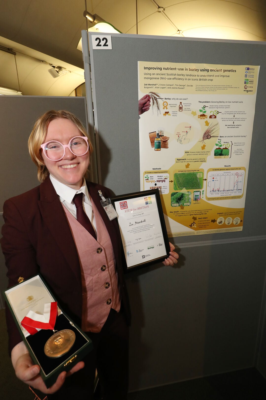 A person smiling while stood next to a poster holding a framed certificate and medal.